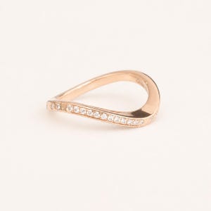 May include: A rose gold ring with a curved design. The ring is adorned with small, sparkling stones set along the outer curve. The ring's smooth, polished surface reflects light, creating a delicate and elegant appearance. A piece of jewelry.