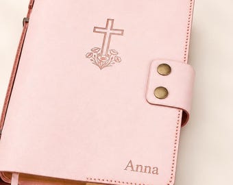 May include: A light pink leather-like journal cover featuring a debossed cross with floral accents at the center and the name Anna engraved in the bottom right corner. The cover includes a side strap with two brass-colored metal snap buttons.