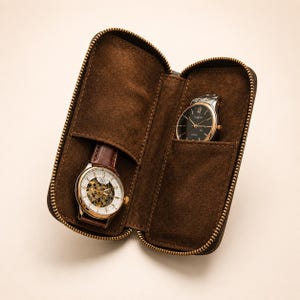 Pode incluir: Um estojo de viagem para relógios em camurça castanho-escura com fecho, contendo dois relógios de pulso. O relógio da esquerda tem um mostrador esqueleto com bracelete de pele castanha, enquanto o da direita tem mostrador preto e bracelete metálica.
