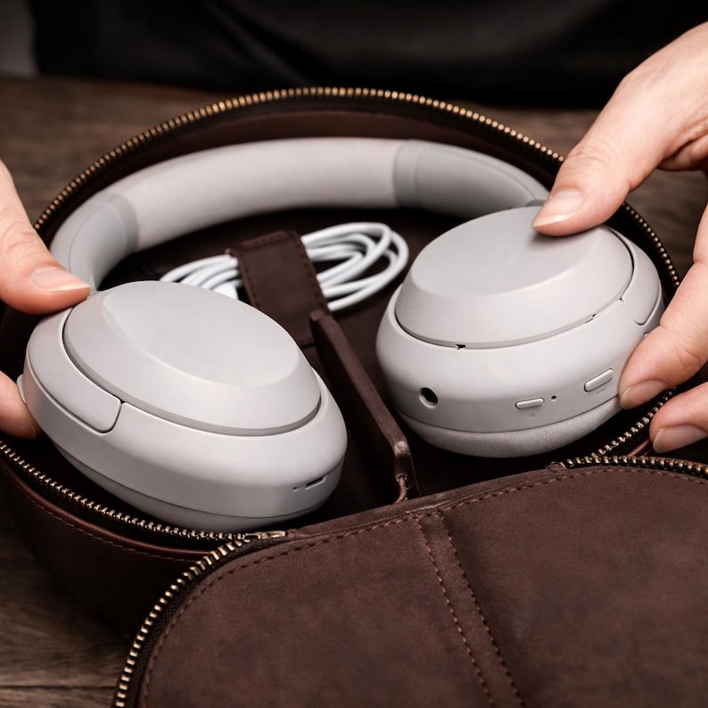 May include: Light grey wireless headphones with a matching headband and white cable, inside a brown leather zippered case. The headphones are being held by a person's hands.