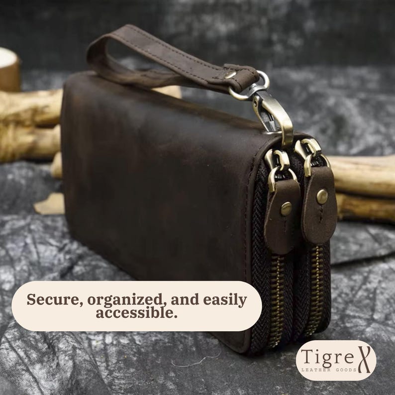 Op de afbeelding: Een donkerbruine leren clutch met een afneembare polsband, twee hoofdvakken met rits en messingkleurige hardware, en een tekstoverlay die luidt: Secure, organized, and easily accessible. Het merklogo luidt: Tigre Leather Goods.
