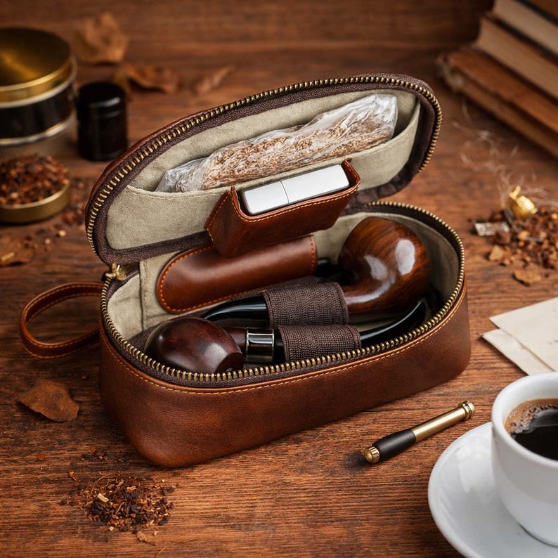 May include: A brown leather pipe case, open to reveal two wooden pipes, a lighter, and tobacco. The case is on a wooden surface with a cup of coffee and a pipe tool nearby. The case has a zipper closure.