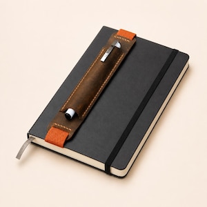 Puede incluir: Un cuaderno negro con cierre elástico y un portalápices de cuero marrón sujeto con correas naranjas. Un bolígrafo plateado está en el soporte. El cuaderno tiene un marcador de cinta gris.