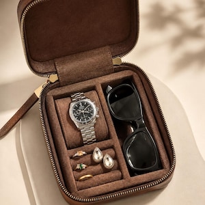 May include: A brown leather travel jewelry case with a zippered closure, containing a silver chronograph watch on a cushion, black sunglasses, two teardrop-shaped earrings, two rings with colored stones, and one plain gold-tone band ring.