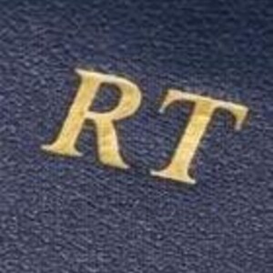 May include: Close-up of a navy blue leather-textured surface with the gold letters "RT". The letters are in a serif font and are slightly raised from the surface. The surface has a subtle, textured pattern.
