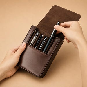 May include: A brown leather pen case, open to reveal several pens. One hand is removing a pen from the case. The case is rectangular with a flap closure. The pens are silver and black.