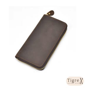 Op de afbeelding: Een donkerbruine leren portemonnee met een gladde, matte afwerking en een goudkleurige metalen ritssluiting die langs drie zijden loopt. De portemonnee heeft een bijpassende leren ritstrekker en bevat de tekst Tigre Leather Goods.