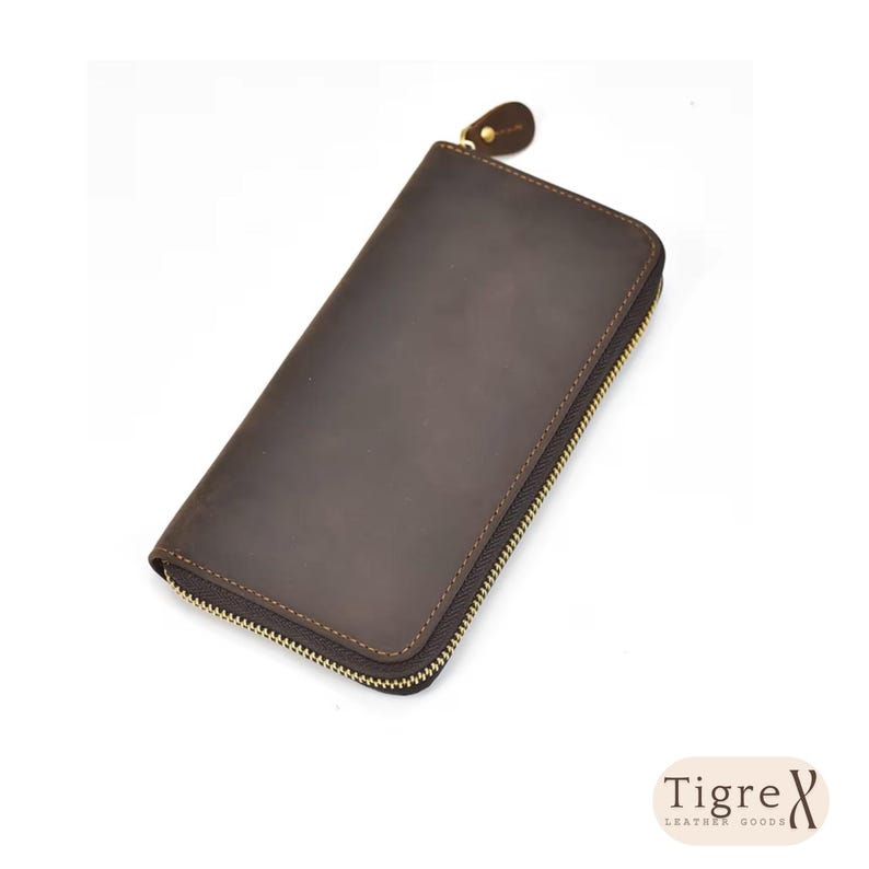 Op de afbeelding: Een donkerbruine leren portemonnee met een gladde, matte afwerking en een goudkleurige metalen ritssluiting die langs drie zijden loopt. De portemonnee heeft een bijpassende leren ritstrekker en bevat de tekst Tigre Leather Goods.