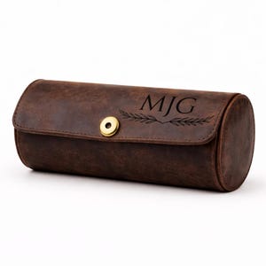 Può includere: Un astuccio per orologi in pelle marrone di forma cilindrica. Presenta una chiusura a patta con un bottone automatico color oro. Le iniziali "MJG" sono incise sul davanti, accompagnate da un motivo decorativo a foglie. La pelle ha un aspetto invecchiato.
