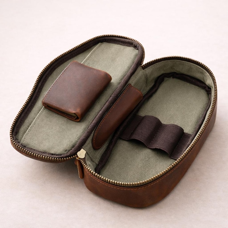 May include: Open brown leather pencil case with a brass zipper. Inside, a small brown leather wallet and a pen holder are visible. The interior is lined with a light green fabric. The case is designed to hold writing instruments and small accessories.