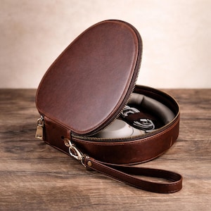 May include: A brown leather headphone case with a zip closure, open to reveal headphones and a cable. The case has a wrist strap and a silver-coloured clasp. The case is on a wooden surface.