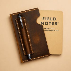 May include: A dark brown leather notebook cover with visible light-colored stitching and a built-in pen holder. A black pen is inserted into the holder. A tan Field Notes memo book with black text is partially tucked inside the leather cover.