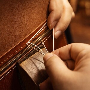 May include: Close-up of a leather item being hand-stitched. The brown leather has a zipper and is being sewn with white thread. A wooden block is used to hold the leather in place during the stitching process.