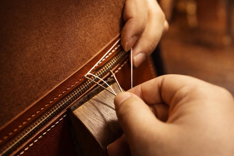 May include: Close-up of a leather item being hand-stitched. The brown leather has a zipper and is being sewn with white thread. A wooden block is used to hold the leather in place during the stitching process.