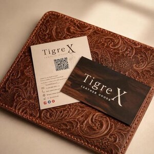 Op de afbeelding: Een vierkante bruine leren mat met een reli&euml;f bloemenpatroon. Bovenop liggen twee rechthoekige visitekaartjes. E&eacute;n kaart is cr&egrave;me met de tekst Tigre X en een QR-code. De andere is donkerbruin met de tekst Tigre X LEATHER GOODS.