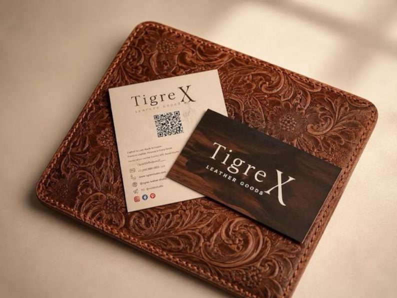 Op de afbeelding: Een vierkante bruine leren mat met een reli&euml;f bloemenpatroon. Bovenop liggen twee rechthoekige visitekaartjes. E&eacute;n kaart is cr&egrave;me met de tekst Tigre X en een QR-code. De andere is donkerbruin met de tekst Tigre X LEATHER GOODS.