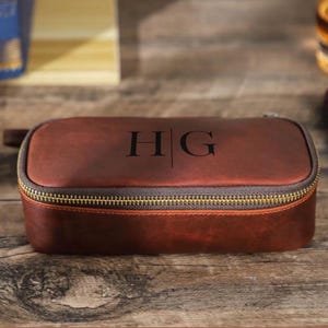 May include: A brown leather travel case with a gold zipper and the initials "HG" printed on the top. The case is rectangular with rounded edges and a small handle on one side. The case is sitting on a wooden surface.