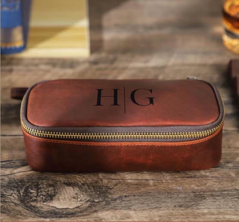 May include: A brown leather travel case with a gold zipper and the initials "HG" printed on the top. The case is rectangular with rounded edges and a small handle on one side. The case is sitting on a wooden surface.