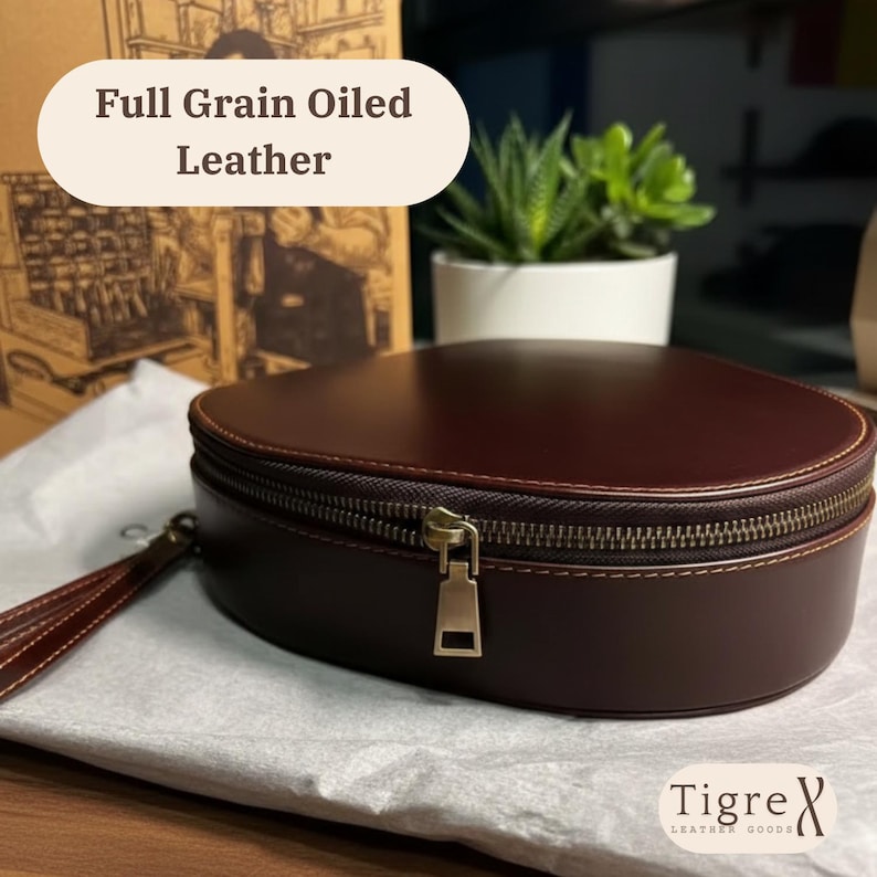 May include: A dark brown, oval leather clutch with a brass zip and a wrist strap. The text "Full Grain Oiled Leather" is displayed above the clutch. The brand name "Tigre X Leather Goods" is visible at the bottom.