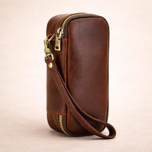May include: A brown leather clutch with a rectangular shape and a zipper closure. The clutch features a wrist strap and a gold-colored metal zipper and clasp. The leather has a distressed appearance.