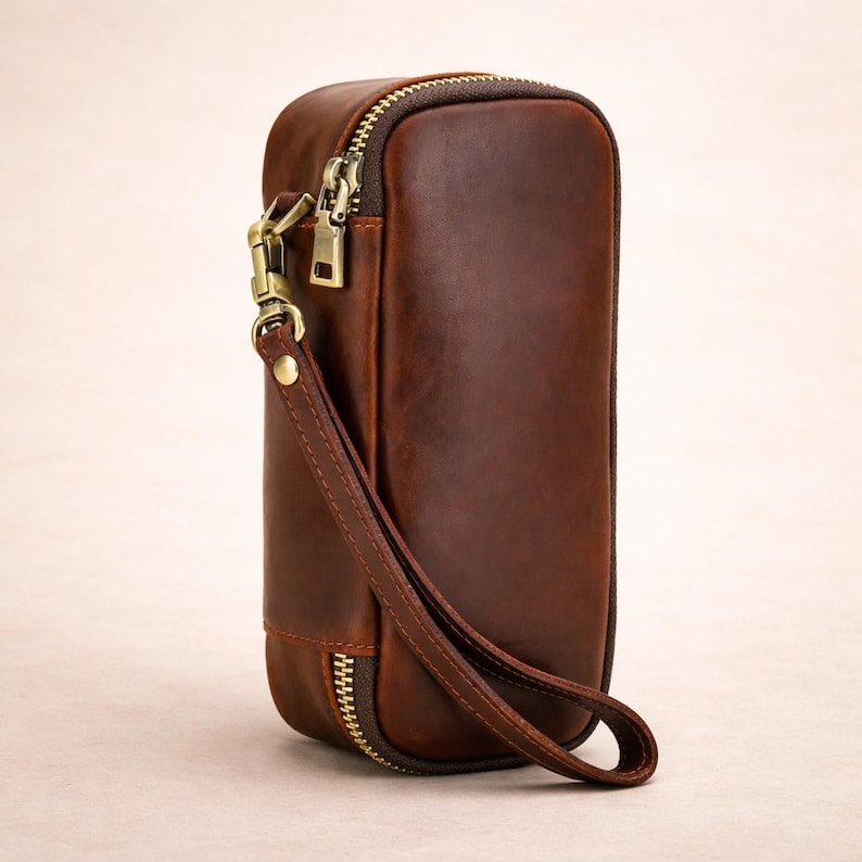 May include: A brown leather clutch with a rectangular shape and a zipper closure. The clutch features a wrist strap and a gold-colored metal zipper and clasp. The leather has a distressed appearance.