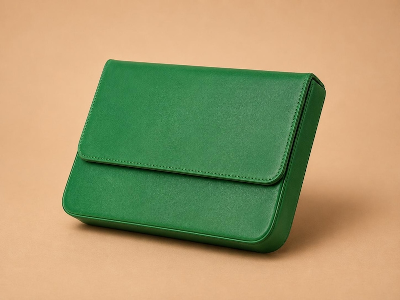 Pode incluir: Uma clutch retangular verde esmeralda com fecho de aba. A bolsa &eacute; feita de um material liso e apresenta uma borda costurada. A clutch est&aacute; sobre um fundo neutro.