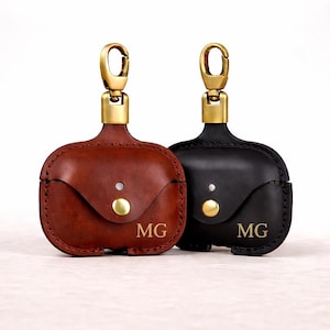 May include: Two leather AirPod cases, one in brown and one in black, with gold-coloured hardware. Each case has a snap closure and the initials "MG" engraved on the front. They have a loop and clasp for attaching to a bag or keyring.