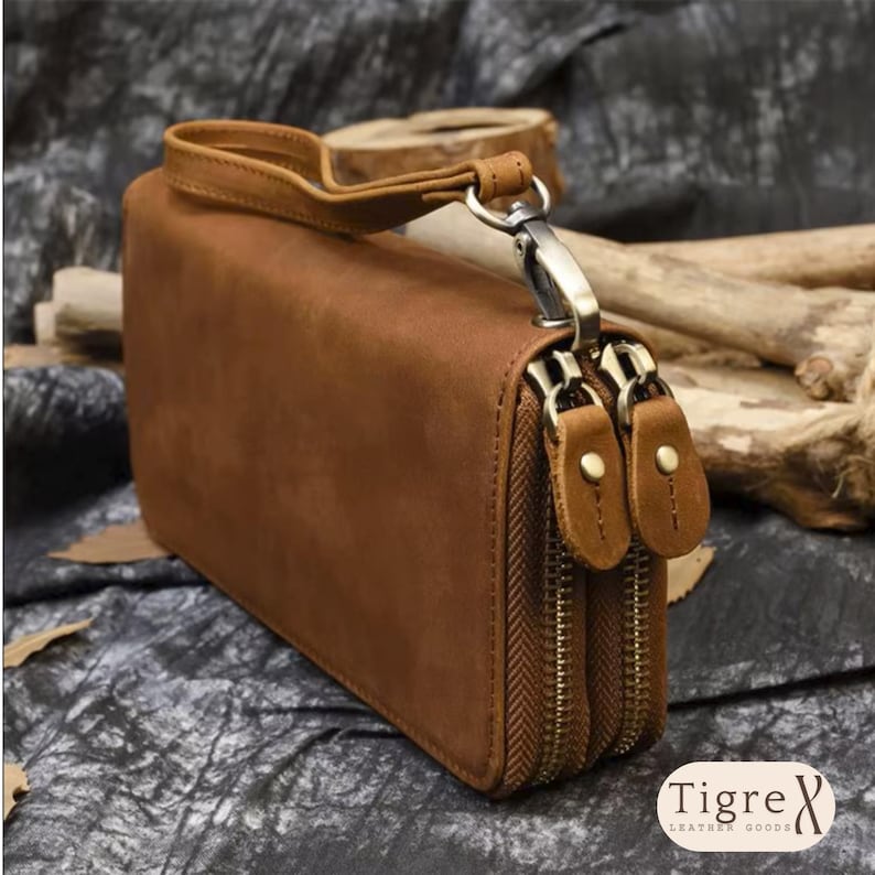 Op de afbeelding: Een beige leren clutch met een afneembare polsband, twee aparte vakken met ritssluiting met bijpassende leren ritstrekkers en een zilverkleurige metalen sluiting. De merknaam Tigre Leather Goods staat in de hoek gedrukt.