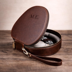 May include: A brown leather travel case with a zip closure and a wrist strap. The case is open, revealing a pair of white shoes and other items inside. The initials "M.E." are embossed on the lid.