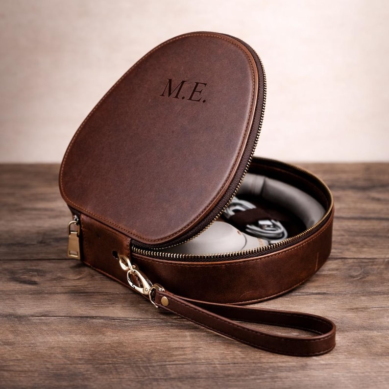 May include: A brown leather travel case with a zip closure and a wrist strap. The case is open, revealing a pair of white shoes and other items inside. The initials "M.E." are embossed on the lid.
