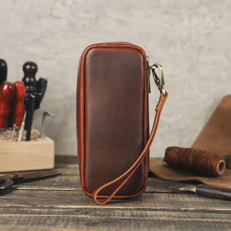 May include: A rectangular, brown leather pouch with a wrist strap. The pouch has a zipper closure and a contrasting reddish-brown trim. The background includes tools and leather crafting supplies, suggesting a handmade product.