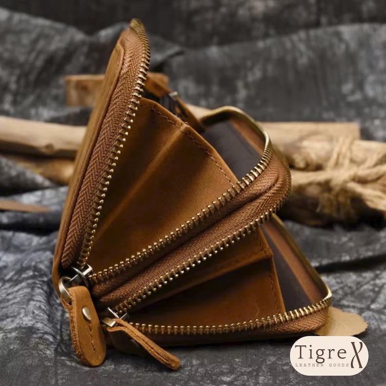 Op de afbeelding: Een bruine lederen portemonnee met een metalen ritssluiting rondom, meerdere interne accordeonvakken en een bijpassende lederen ritstrekker. De merknaam Tigre Leather Goods staat in de rechterbenedenhoek afgedrukt.