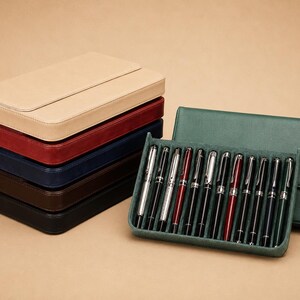 Pode incluir: Uma pilha de estojos de caneta de couro em bege, vermelho, azul marinho e castanho. Uma capa de caneta verde e uma bandeja de canetas prateadas e pretas com detalhes vermelhos tamb&eacute;m est&atilde;o presentes. As canetas s&atilde;o exibidas em uma superf&iacute;cie verde aveludada.