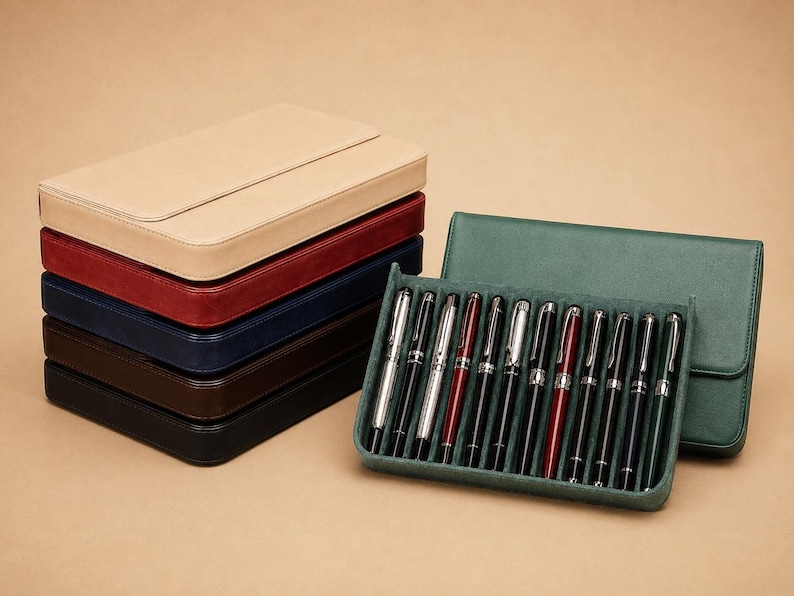 Pode incluir: Uma pilha de estojos de caneta de couro em bege, vermelho, azul marinho e castanho. Uma capa de caneta verde e uma bandeja de canetas prateadas e pretas com detalhes vermelhos tamb&eacute;m est&atilde;o presentes. As canetas s&atilde;o exibidas em uma superf&iacute;cie verde aveludada.