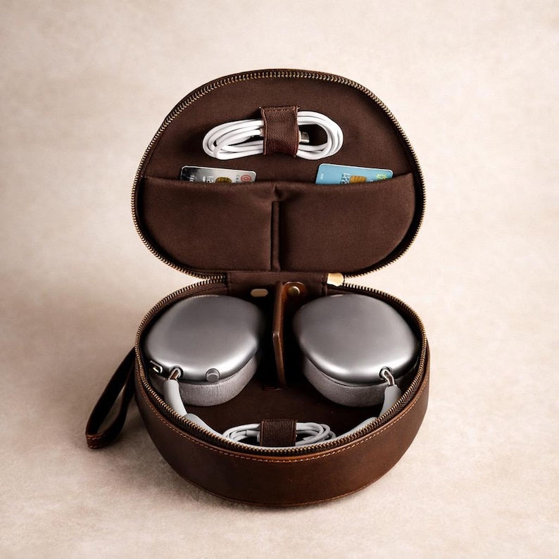 May include: Open brown leather headphone case with a zip, holding silver headphones, a white charging cable, and two small pockets. The case is round with a handle. The interior is a lighter brown.