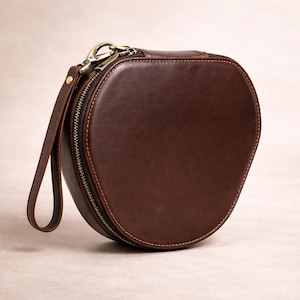 May include: A brown leather, oval-shaped case with a zip closure and a wrist strap. The case has a brass-coloured zip and hardware. The leather appears smooth and has a slight sheen. The case is designed to carry small items.