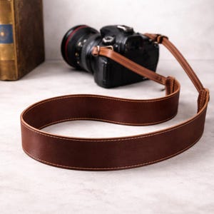 May include: A brown leather camera strap with white stitching is looped in the foreground. A black camera with a lens is in the background. The strap is made of a smooth, durable leather material.
