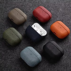 May include: A selection of leather AirPod cases in shades like olive green, navy blue, and orange. One case is open, displaying white AirPods. The cases have a textured finish and a simple design.