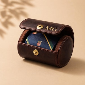 May include: A brown leather tie case with the initials "MG" embossed on the lid. Inside, a rolled-up blue tie with a striped pattern and crest details is visible. The case is open, revealing the tie.