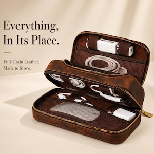 Könnte beinhalten: Geöffnete braune Leder-Reise-Organizer mit Reißverschluss, gefüllt mit weißen elektronischen Zubehör. Der Text "Everything, In Its Place" und "Full-Grain Leather. Made to Move" sind sichtbar. Für Kabel, Ladegeräte und mehr.