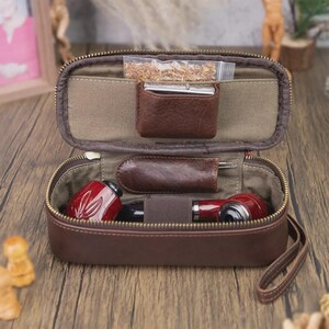 May include: Open brown leather tobacco pipe case with a zipper. Inside, there are two red and black pipes, a small leather pouch, and a clear bag of tobacco. The case is open, revealing the contents.