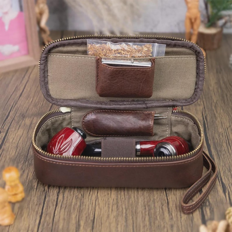 May include: Open brown leather tobacco pipe case with a zipper. Inside, there are two red and black pipes, a small leather pouch, and a clear bag of tobacco. The case is open, revealing the contents.