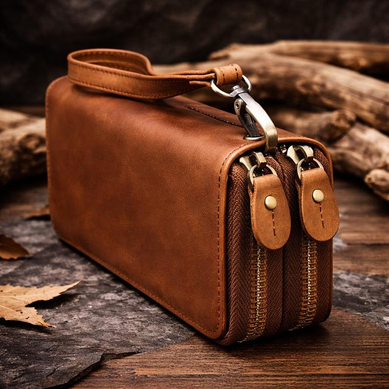 Op de afbeelding: Een rechthoekige bruine leren clutch met een afneembare polsband, voorzien van twee aparte vakken met rits en messingkleurige hardware, zichtbare stiksels langs de randen en twee bijpassende leren ritstrekkers op elke metalen rits.