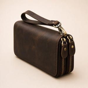 Op de afbeelding: Een donkerbruine leren clutch met een afneembare polsband, twee aparte vakken met messingkleurige metalen ritsen en bijpassende leren ritstrekkers met kleine messing klinknagels op elk treklipje.