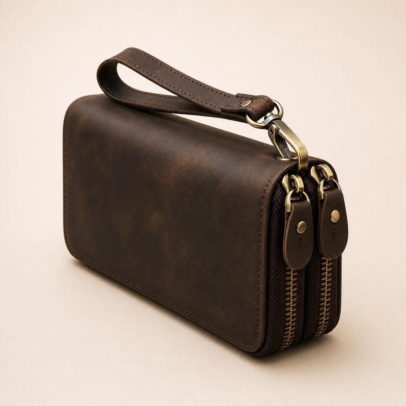 Op de afbeelding: Een donkerbruine leren clutch met een afneembare polsband, twee aparte vakken met messingkleurige metalen ritsen en bijpassende leren ritstrekkers met kleine messing klinknagels op elk treklipje.