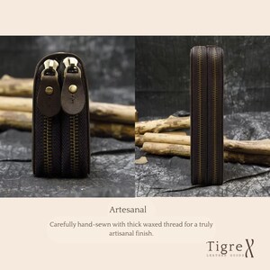Op de afbeelding: Een donkerbruin lederen etui met twee aparte vakken met rits, voorzien van messingkleurige metalen ritsen en bijpassende lederen treklipjes. Het item is met de hand genaaid met dik gewaxt garen en toont de tekst Artesanal en Tigre Leather Goods.