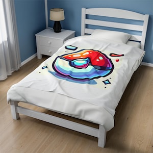 May include: A white fleece blanket with a colorful graphic of a Pokeball. The Pokeball is red, blue, and white with a black outline. The background is a light yellow with white stars.