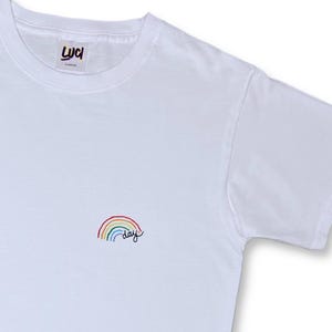 Può includere: T-shirt bianco con un piccolo arcobaleno ricamato e la parola "day" in corsivo. La maglietta ha un girocollo e maniche corte. Il logo del marchio è in alto a sinistra.