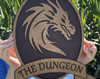 May include: Wooden sign with a dragon head design inside an oval, with a banner below that reads "THE DUNGEON". The sign is made of two tones of wood, with a dark background and a lighter dragon head.