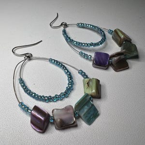 Puede incluir: Un par de pendientes colgantes con diseño de doble aro. El aro interior está bordeado con pequeñas cuentas azul claro translúcidas. El aro exterior está decorado con una serie de cuentas irregulares similares a piedras en tonos morado, verde y gris.